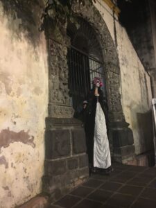 Person dressed as La Catrina standing by an old stone archway, wearing a traditional white dress and floral headdress