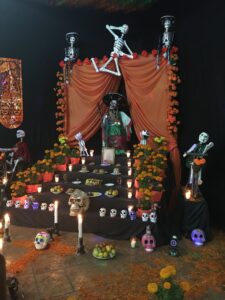 El Día de Muertos [Day of the Dead] altar decorated with skeletons, marigold flowers, candles, sugar skulls, and various offerings.