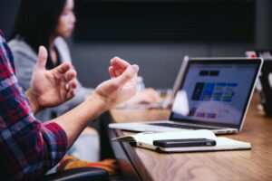 A close-up of a person's hands gesturing during a discussion, with a laptop, notebook, and smartphone on the table in focus, while another participant is blurred in the background