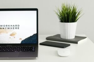 A laptop displaying the text "Work Hard Anywhere" on a minimalistic desk setup, with a smartphone, a notebook, a mouse, and a potted plant.