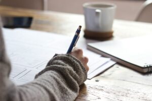 Close-up of a person writing on paper at a wooden desk, with a notebook and coffee mug nearby.