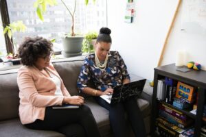 Two women sitting on a couch in an office, one using a laptop and the other holding a notebook