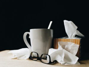 A white mug with a spoon, a box of tissues, and a pair of glasses on a table, symbolizing sickness and the need for rest