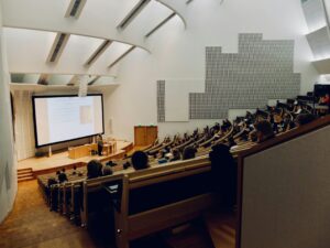 A large, modern lecture hall with students seated in tiered rows, attentively listening to a professor giving a presentation at the front. A large projector screen displays a slide with text and an image