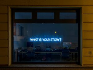  A neon sign in an office window displaying the words "What Is Your Story?"