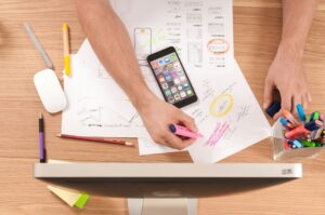 A workspace with hands highlighting notes, a smartphone, and various pens spread across a desk