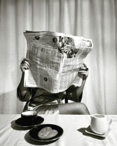 A person sitting at a table reading a newspaper, with a cup of coffee and cookies in front of them.