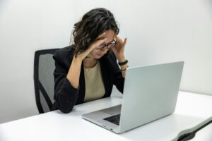 A woman wearing glasses and a black blazer sits at a white desk with a silver laptop. She holds her head with both hands, appearing stressed or deep in thought.