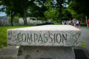 A stone bench with the word "COMPASSION" engraved on it, set in an outdoor park with trees and a group of people in the blurred background.