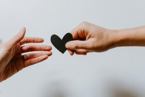 Two hands reaching out to each other, one giving a small black heart-shaped paper cutout to the other