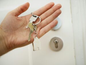 A hand holding a set of keys with a small house-shaped keychain in front of a door lock.